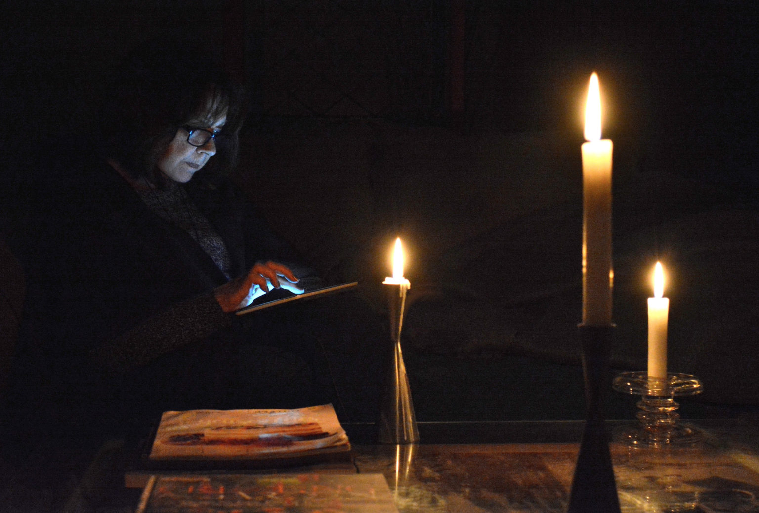 woman reading by candle light on her digital tablet, while awaiting the return of power to her home.